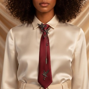 Peut inclure: Une cravate bordeaux avec un fermoir décoratif en métal orné d'oiseaux et de plumes. La cravate est portée avec une chemise boutonnée crème et un pantalon beige. Le fermoir en métal a des perles pendantes et une chaîne.