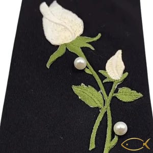 Pu&ograve; includere: Tessuto nero con una rosa e un bocciolo ricamati in bianco, con foglie e stelo verdi. Due perle sono posizionate lungo lo stelo. Un logo di pesce dorato si trova nell'angolo in basso a destra.