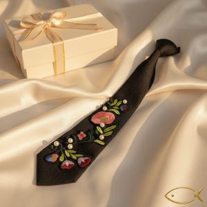 Puede incluir: Una corbata negra con detalles florales bordados en rosa, rojo y verde, acentuada con peque&ntilde;as perlas blancas. Una caja de regalo con una cinta dorada se encuentra en el fondo. Accesorio formal.