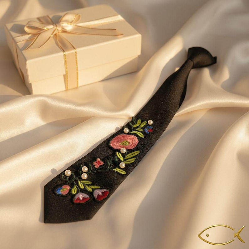 Puede incluir: Una corbata negra con detalles florales bordados en rosa, rojo y verde, acentuada con peque&ntilde;as perlas blancas. Una caja de regalo con una cinta dorada se encuentra en el fondo. Accesorio formal.