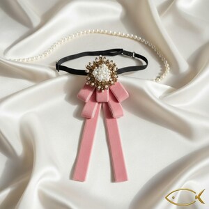 K&ouml;nnte beinhalten: Eine rosa Samt-Fliege mit einer dekorativen Gold- und Perlenverzierung. Die Fliege ist an einem schwarzen Gummiband befestigt. Eine Perlenkette liegt auf einer wei&szlig;en Satin-Oberfl&auml;che.
