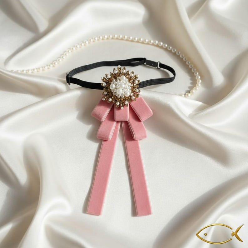 K&ouml;nnte beinhalten: Eine rosa Samt-Fliege mit einer dekorativen Gold- und Perlenverzierung. Die Fliege ist an einem schwarzen Gummiband befestigt. Eine Perlenkette liegt auf einer wei&szlig;en Satin-Oberfl&auml;che.
