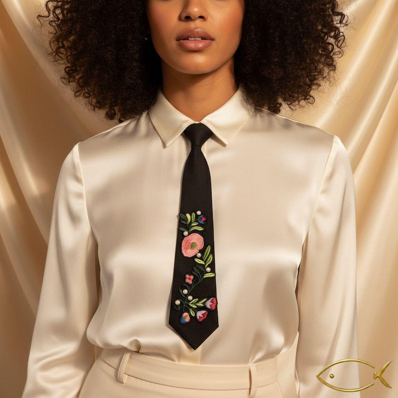 Puede incluir: Una corbata negra con dise&ntilde;o floral bordado, con flores rosas, rojas y azules con hojas verdes y detalles de perlas blancas. La corbata se lleva con una camisa blanca con cuello y pantalones beiges.