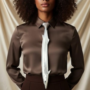 Può includere: Una cravatta argentata a forma di pesce, indossata con una camicia di raso marrone e pantaloni marroni. La cravatta presenta dettagli come pinne e testa. La camicia ha un colletto classico e maniche lunghe.
