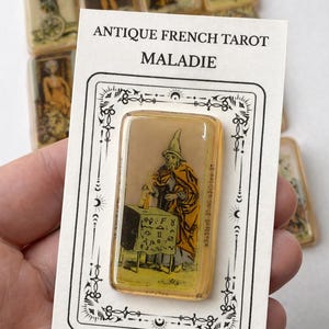 Może przedstawiać: Ręcznie robiona płytka ze stopionego szkła z wizerunkiem z antycznej francuskiej karty tarota. Karta przedstawia postać w pomarańczowej szacie i kapeluszu, stojącą za stołem. Widoczny jest tekst "MALADIE".