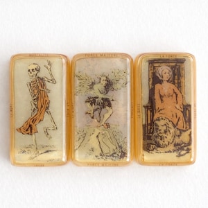 May include: Three vintage tarot cards with gold-colored frames. The cards feature illustrations of a skeleton, an angel, and a woman with a lion. The cards have French text, including "Mortalite", "Force Majeure", and "La Force".