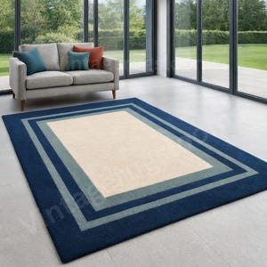 May include: A rectangular area rug with a navy blue border, a light blue inner border, and a cream-colored center. The rug is placed on a tiled floor in a well-lit room with a sofa and large windows.
