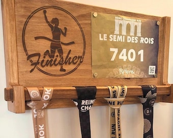 May include: A rectangular wooden wall-mounted display rack featuring a laser-engraved runner silhouette and the word Finisher. A race bib labeled LE SEMI DES ROIS 7401 is attached. Four race medals hang from the rack by their branded ribbons.
