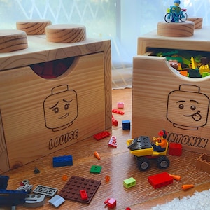 Może przedstawiać: Dwa drewniane pudełka do przechowywania zabawek, zaprojektowane tak, aby przypominały duże klocki. Każde pudełko ma szufladę z twarzą w stylu Lego i imieniem. Jedno pudełko jest otwarte, ukazując kolorowe zabawki.