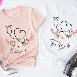 Könnte beinhalten: Zwei T-Shirts, eines in Heather Peach und eines in Weiß, mit einem Stethoskop-Design. Das Pfirsichfarbene sagt "Bridesmaids" und das weiße Shirt sagt "The Bride". Beide Shirts haben Blumendetails und die Sätze "She said yes" oder "I said yes".