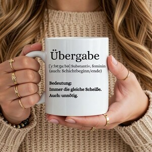 Könnte beinhalten: Weißer Keramikbecher mit dem deutschen Wort "Übergabe" aufgedruckt, zusammen mit seiner Definition und Bedeutung. Der Becher wird von einer Person gehalten, die goldene Ringe und einen beigen Pullover trägt.
