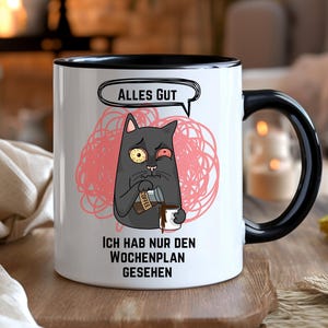 Könnte beinhalten: Weiße Keramiktasse mit schwarzem Henkel und Rand. Die Tasse zeigt eine Cartoon-Katze mit müdem Gesichtsausdruck, eine Sprechblase mit den Worten "ALLES GUT" und dem Text "ICH HAB NUR DEN WOCHENPLAN GESEHEN". Die Katze gießt Kaffee in eine Tasse.