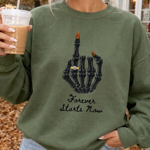 Könnte beinhalten: Ein olivgrünes Sweatshirt mit Rundhalsausschnitt und einer Grafik einer schwarzen Skeletthand, die den Mittelfinger zeigt. Die Hand trägt einen goldenen Ring mit einem weißen Stein. Unter der Grafik steht der Text Forever Starts Now in schwarzer Schrift.
