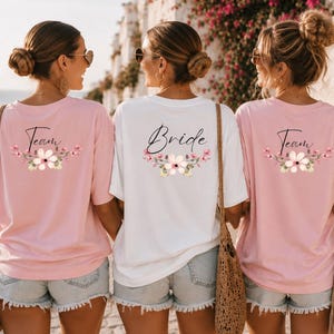 Könnte beinhalten: Drei Frauen tragen passende T-Shirts. Zwei rosa Shirts mit der Aufschrift "Team" und einem Blumendesign. Das weiße Shirt trägt die Aufschrift "Bride" mit einem Blumendesign. Alle Shirts sind kurzärmlig.