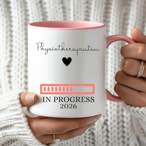 Könnte beinhalten: Weißer Keramikbecher mit rosa Innenraum und Henkel. Der Becher trägt den Text "Physiotherapeutin" über einem schwarzen Herzen, einen Fortschrittsbalken und "IN PROGRESS 2026". Der Becher wird von einer Person gehalten, die einen weißen Pullover trägt.