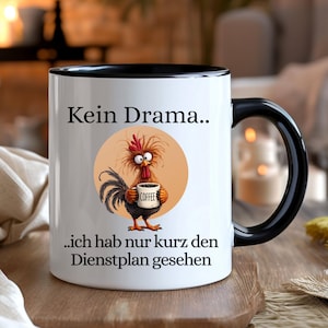 May include: White ceramic coffee mug with a black handle and rim. The mug features a cartoon rooster holding a coffee cup, with the text "Kein Drama.. ich hab nur kurz den Dienstplan gesehen."