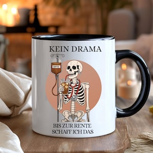 Könnte beinhalten: Weiße Keramik-Kaffeetasse mit schwarzem Henkel und Rand. Die Tasse zeigt eine Skelett-Illustration mit Stethoskop, das einen Kaffeebecher hält. Auf der Tasse steht "KEIN DRAMA" und "BIS ZUR RENTE SCHAFF ICH DAS."
