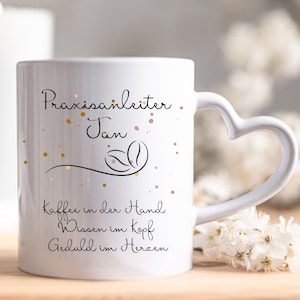 Op de afbeelding: Witte keramische mok met een hartvormig handvat. De mok heeft de tekst "Praxisanleiter Jan" en een koffieboonontwerp, samen met extra tekst in het Duits. Goudkleurige spikkels voegen een decoratief tintje toe. De mok staat op een houten oppervlak.