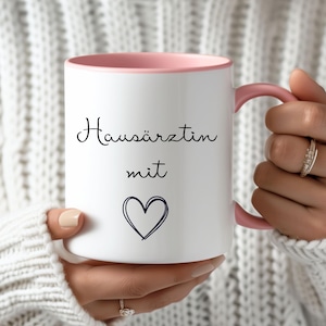 Puede incluir: Taza de cerámica blanca con interior y asa rosa. La taza presenta las palabras "Hausärztin mit" en escritura negra, junto con un corazón azul dibujado a mano. La taza es sostenida por una persona que lleva un suéter blanco.