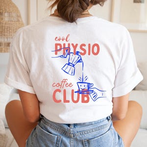 May include: A white t-shirt featuring a graphic on the back with the text "cool PHYSIO coffee CLUB" in coral red. The design includes blue line art of a hand pouring coffee from a moka pot into a smiling cup held by another hand.