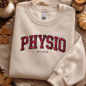 May include: A beige crewneck sweatshirt featuring the word PHYSIO in a red and black plaid pattern across the chest. Below the word, the text EST.2026 is printed in a simple black font. The sweatshirt has long sleeves and a ribbed collar.