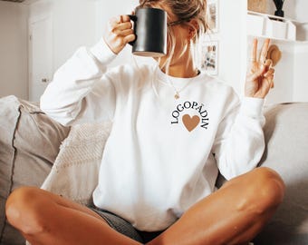 May include: White sweatshirt with the word "LOGOPÄDIN" arched above a brown heart. The person is holding a black mug. The person is wearing a gold necklace and fuzzy slippers.