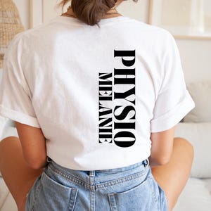 Puede incluir: Una camiseta blanca con las palabras "PHYSIO MELANIE" impresas verticalmente en negro. La camiseta está hecha de un material suave y se está usando.