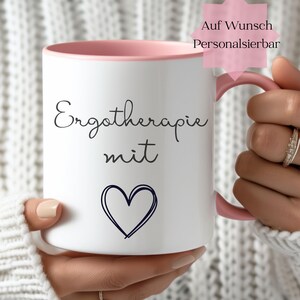 Könnte beinhalten: Weißer Keramikbecher mit rosa Innenraum und Henkel. Der Becher trägt die Aufschrift "Ergotherapie mit" in schwarzer Schrift sowie ein handgezeichnetes Herz. Der Text "Auf Wunsch Personalisierbar" befindet sich auf einem rosa sternförmigen Etikett.