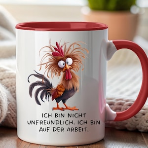 Könnte beinhalten: Ein weißer Keramikbecher mit rotem Innenraum und Henkel, verziert mit einer Cartoon-Illustration eines zerzausten braunen Hahns mit großen Augen. Darunter steht der schwarze Text: ICH BIN NICHT UNFREUNDLICH. ICH BIN AUF DER ARBEIT.