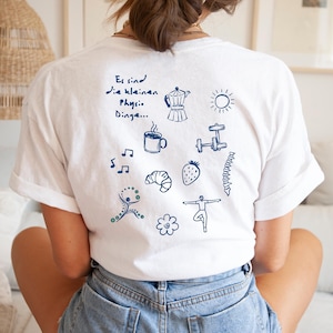 Könnte beinhalten: Ein weißes T-Shirt mit dunkelblauen Strichzeichnungen auf der Rückseite, darunter eine Kaffeekanne, Sonne, Tasse, Hantel, Noten, Croissant, Erdbeere, Wirbelsäule, Blume und Figuren, mit dem Text Es sind die kleinen Physio Dinge.