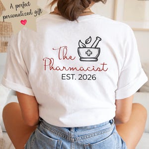 Könnte beinhalten: Weißes T-Shirt mit dem Text "The Pharmacist" in roter Schrift und einer schwarzen Mörser- und Stößel-Grafik. Darunter steht "EST. 2026". Die Person trägt blaue Jeansshorts.