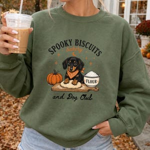 Könnte beinhalten: Ein olivgrüner Rundhals-Pullover mit einer Grafik eines schwarz-braunen Dackels mit orangefarbenem Halstuch, der auf einem Holzbrett Teig knetet, neben einem Kürbis und einer Schüssel mit der Aufschrift FLOUR sowie dem Text SPOOKY BISCUITS bakery and Dog Club.