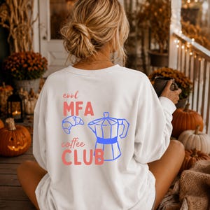 Puede incluir: Una sudadera blanca de manga larga con un gráfico en la espalda que incluye el texto "cool MFA coffee CLUB" en color rosa. El diseño presenta el contorno azul de un cruasán y el contorno azul de una cafetera moka centrados en la espalda.