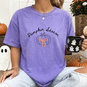May include: A purple short-sleeve t-shirt featuring the black text Pumpkin Season above a black line-art pumpkin graphic with an orange bow. The shirt has a crew neckline and a relaxed fit, showcasing a casual style for the autumn season.