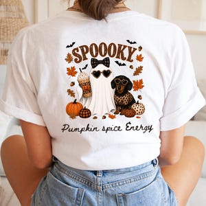 Könnte beinhalten: Ein weißes T-Shirt mit einem Rückendruck, der den Text SPOOOKY und Pumpkin spice Energy zeigt. Das Motiv umfasst ein Gespenst mit Schleife und herzförmiger Sonnenbrille, einen Dackel im Leopardenpullover, Kürbisse und Herbstblätter.