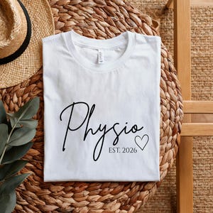 May include: A white short-sleeved t-shirt featuring black text that reads Physio in a cursive script, followed by the text EST. 2026 and a small black heart outline. The shirt has a crew neckline and a standard fit, presented in a flat layout.