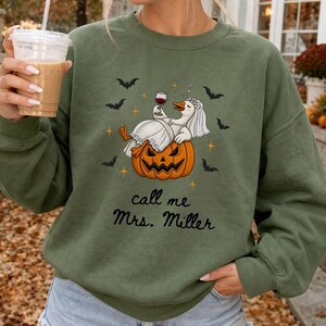 May include: An olive green crewneck sweatshirt featuring a graphic of a white goose wearing a bridal veil and necklace, lounging on a carved orange jack-o'-lantern while holding a glass of red wine, surrounded by black bats and the text call me Mrs. Miller.