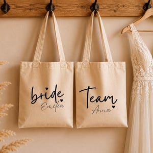 Könnte beinhalten: Zwei beigefarbene Tragetaschen mit schwarzer Schrift, die "bride Emilia" und "Team Anna" lautet. Die Taschen hängen an einer Holzleiste, rechts daneben ein Brautkleid auf einem Bügel. Die Taschen haben lange Henkel.