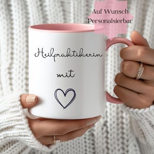 Könnte beinhalten: Weißer Keramikbecher mit rosa Innenraum und Henkel. Der Becher trägt die Worte "Heilpraktikerin mit" in schwarzer Schrift sowie ein handgezeichnetes blaues Herz. Ein rosa sternförmiges Etikett lautet "Auf Wunsch Personalisierbar".