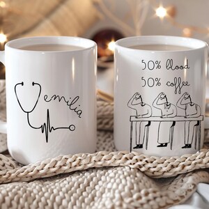 Könnte beinhalten: Zwei weiße Keramikbecher. Ein Becher zeigt eine Stethoskop-Grafik, den Namen "Emilia" und eine Herzschlaglinie. Der andere Becher hat den Text "50% blood 50% coffee" und eine Strichzeichnung von Personen.