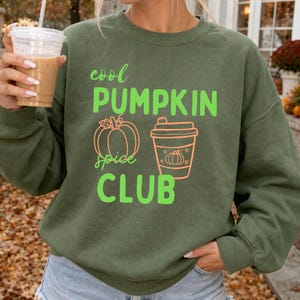 Könnte beinhalten: Ein olivgrüner Rundhals-Pullover mit dem Schriftzug "cool PUMPKIN spice CLUB" in hellem Limettengrün. Unter dem Text befinden sich Strichzeichnungen eines Kürbisses und eines Kaffeebechers in hellem Orange, wobei der Becher kleine Herzen zeigt.