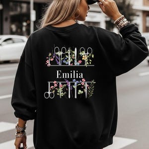 Può includere: Felpa nera con un design floreale e forbici. La parola "Emilia" è stampata in bianco. Il design include forbici, fiori e verde. La felpa è a girocollo e maniche lunghe.