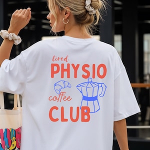 May include: White t-shirt featuring the text "tired PHYSIO coffee CLUB" in orange and blue, with a croissant and coffee pot graphic. The shirt has a relaxed, oversized style.
