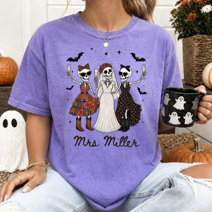 May include: A purple t-shirt featuring a graphic of three skeletons holding champagne glasses. The center skeleton wears a white wedding dress and veil, flanked by two skeletons in patterned dresses and cowboy boots. The text Mrs Miller is printed below.