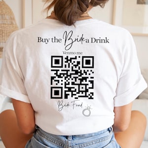 Puede incluir: Camiseta blanca con el texto "Buy the Bride a Drink" y un código QR para Venmo. Debajo del código QR está el texto "Bride Fund" y un gráfico de anillo. La camiseta es de estilo casual.