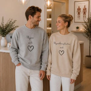 May include: A light gray sweatshirt with the text Physio mit and a heart icon, and a beige sweatshirt with the text Physiotherapeutin mit and a heart icon. Both garments feature long sleeves, crew necklines, and a simple, clean design.