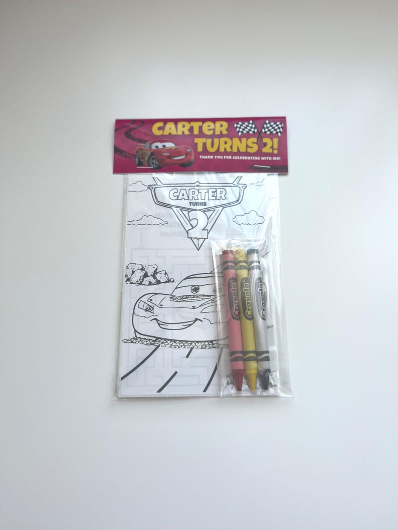 Puede incluir: Un juego de recuerdos para fiestas con un libro para colorear y crayones. El libro para colorear tiene un tema de coches de carreras con el texto "CARTER TURNS 2!". El juego incluye cuatro crayones rojos, rosas, amarillos y grises. El embalaje es de pl&aacute;stico transparente.