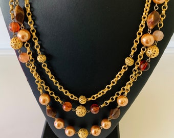 May include: A three-strand necklace with gold-tone chains and various beads. The necklace features large faux pearls, amber-colored beads, and filigree accents. The necklace is displayed on a black jewelry stand.