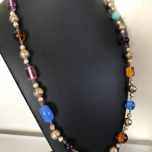 May include: A long necklace with silver beads and colorful glass beads. The beads are in various shapes and sizes, including round, oval, and rectangular. The necklace is on a black display stand.