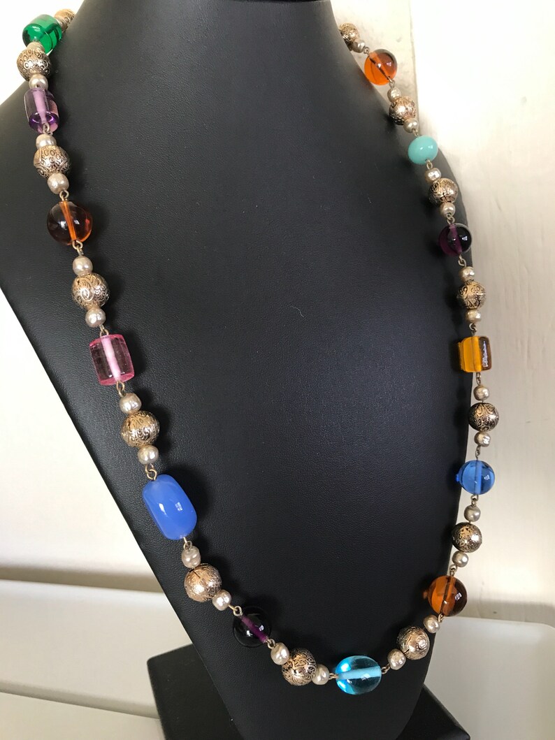 May include: A long necklace with silver beads and colorful glass beads. The beads are in various shapes and sizes, including round, oval, and rectangular. The necklace is on a black display stand.